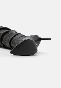 Schwarze Lederstiefel mit hohen Absätzen, spitz zulaufender Zehenpartie und strukturiertem Sohlenprofil. Sie verfügen über geraffte Details entlang des Schafts und einen dünnen Stiletto-Absatz.