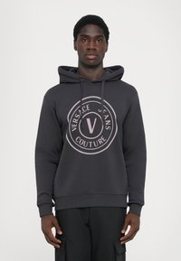 Tmavohnedá mikina s klokankou, ktorá má veľké ružové kruhové logo s textom "VERSACE JEANS COUTURE". Mäkká látka s rebrovanými manžetami.