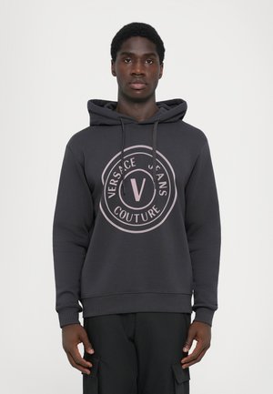 Sudadera gris oscura con un bolsillo tipo canguro, que presenta un gran logo circular rosa con el texto "VERSACE JEANS COUTURE". Tejido suave con puños acanalados.