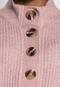 Gros plan d'un pull en maille côtelée rose avec quatre grandes boutons ronds en écaille de tortue sur le devant.