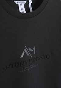 T-shirt nera a maniche corte con collo a giro. Presenta un logo in rilievo e testo in grigio scuro, realizzato in morbido cotone.