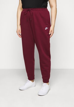 Person iført bordeaux Nike sweatpants med snor og hvide sneakers, stående mod en ensfarvet hvid baggrund.