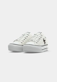 Witte low-top sneakers met dikke rubberen zolen, veters, zwarte rand en een klein cartoon gezicht embleem aan de buitenzijde.
