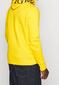 Felpa gialla con cappuccio in tessuto morbido, con polsini e orlo a coste, con testo nero all'interno del cappuccio. Indossata con jeans blu.