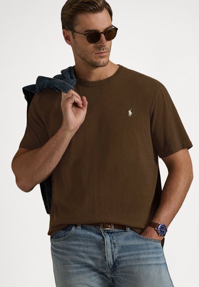 Κοντομάνικο καφέ μπλουζάκι με στρογγυλή λαιμόκοψη και μικρό λευκό λογότυπο στο αριστερό στήθος· συνοδευόμενο από ανοιχτό μπλε τζιν παντελόνι και γυαλιά ηλίου.