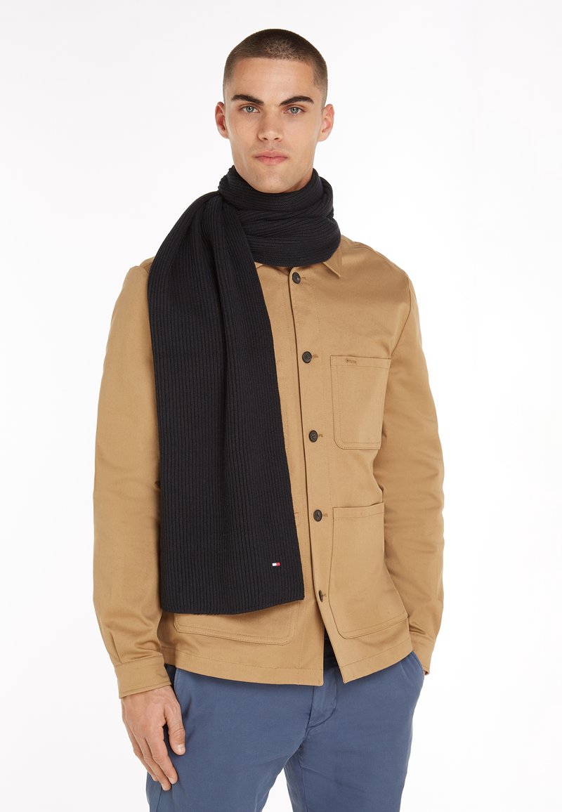 Mann trägt eine hellbraune, geknöpfte Jacke, dunkelblaue Hose und einen langen schwarzen Schal, der um den Hals gewickelt ist.