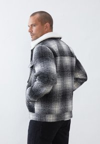 Giacca a scacchi bianca e nera con colletto in pelliccia sintetica, caratterizzata da un tessuto testurizzato e due tasche laterali, abbinata a jeans neri.