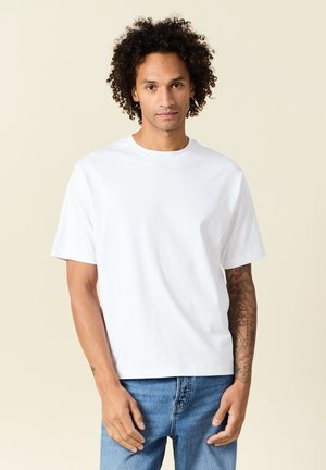 T-shirt bianca in cotone con maniche corte, scollatura a girocollo e vestibilità comoda, esposta su uno sfondo neutro. Indossati jeans in denim blu.