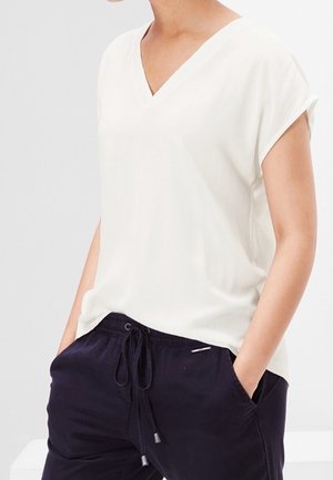 Persoon draagt een wit T-shirt met korte mouwen en V-hals en zwarte broek met trekkoord, met handen in de zakken, staand tegen een effen achtergrond.