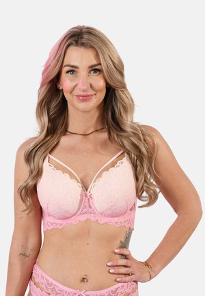 Woman with long blonde hair wearing pink lace bra and matching underwear, standing with hand on hip against light background.