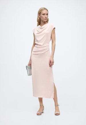 Kvinde iført en lys rosa ærmeløs midi-kjole med draperet halsudskæring, der holder en sølv clutch og har sølv åbne tå-sandaler på.