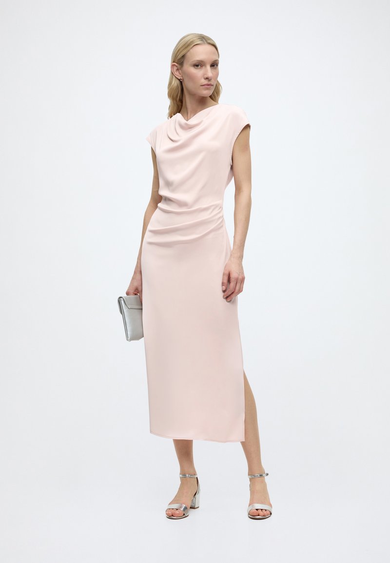 Femme portant une robe midi sans manches rose pâle avec un décolleté drapé, tenant une pochette argentée et portant des talons ouverts argentés.