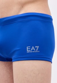 Nærbilde av en person som har på seg tettsittende, kongeblå badeshorts med hvit EA7 Emporio Armani-logo på venstre side.
