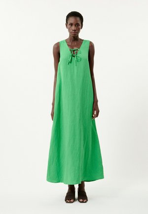 Robe maxi en lin vert avec un décolleté en V et un détail à lacets à l'avant. Texture lisse et coupe décontractée, associée à des sandales en cuir brun foncé.