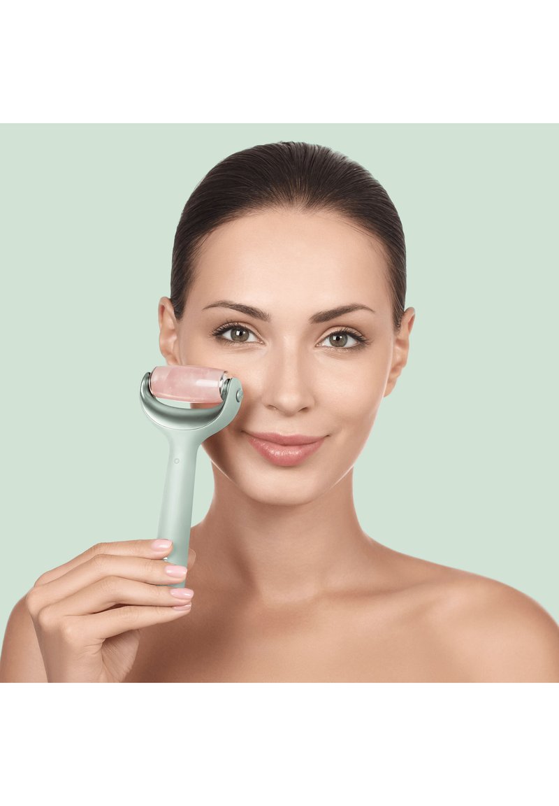 Femme utilisant un rouleau facial en quartz rose sur sa joue, peau claire, cheveux lissés en arrière, fond neutre.