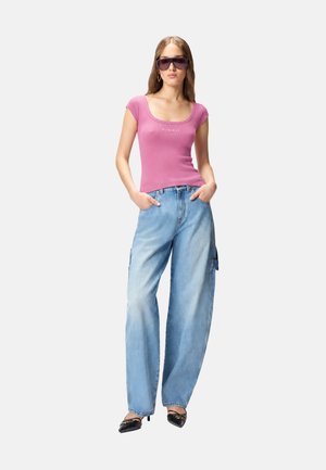 Mujer que lleva vaqueros azules claros de gran tamaño, camiseta rosa ajustada de manga corta, tacones negros puntiagudos y grandes gafas de sol oscuras, de pie con las manos en los bolsillos.