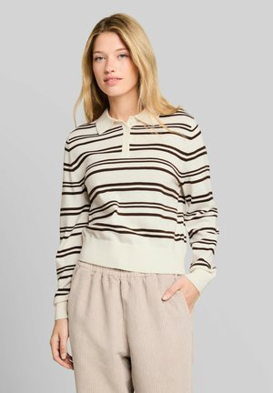 Blonde vrouw die een crème en bruin gestreepte lange mouwen polo draagt en lichtbeige broek, met één hand in haar zak.
