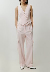 Helles rosa, tailliertes Weste mit V-Ausschnitt und fünf goldenen Knöpfen, kombiniert mit weiten, hoch taillierten Hosen mit Seitentaschen. Weiße spitze Schuhe.