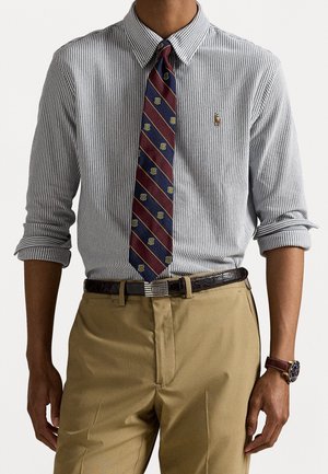 Man wearing a striped button-up shirt with a patterned tie, beige pants with a black belt, and a wristwatch on left wrist.