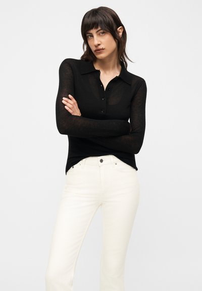 Γυναίκα με σκούρα μαλλιά και αφέλειες, που φοράει διαφανές μαύρο μακρυμάνικο πόλο και λευκό παντελόνι ψηλόμεσο, με σταυρωμένα χέρια, ουδέτερο φόντο.