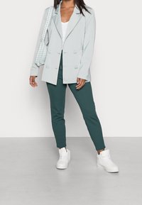 Ljusblå dubbelknäppt kavaj, vit topp, gröna smala byxor och vita sneakers. Kavajen har stora knappar och en avslappnad passform.