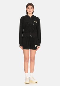 Felpa nera in velluto con zip e tasca frontale, con la scritta "Juicy" in giallo. Indossata con pantaloni corti neri abbinati. Completata con calzini bianchi e sneaker.