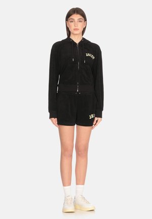 Felpa nera in velluto con zip e tasca frontale, con la scritta "Juicy" in giallo. Indossata con pantaloni corti neri abbinati. Completata con calzini bianchi e sneaker.