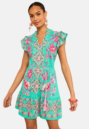 Femme portant une robe bleu sarcelle avec des motifs floraux et géométriques roses, des manches courtes volantées et une ceinture à pompons rose, accessoirisée de boucles d'oreilles et d'un bracelet à fleurs.