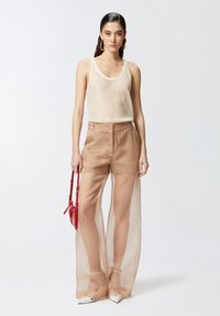 Bež čipkaste široke hlače s visokim strukom, uparene s laganom, strukturiranom majicom bez rukava; model drži malu crvenu torbicu i nosi bijele cipele.