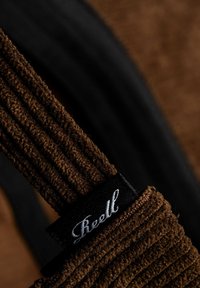 Correa de tela acanalada marrón con un acento negro y una etiqueta de logo blanca. Textura rica con fibras visibles, en contraste con un fondo negro suave.
