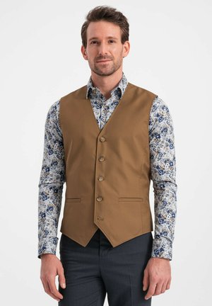 Chaleco marrón ajustado con un dobladillo en forma de V y seis botones, usado sobre una camisa azul con estampado floral. Tejido suave y diseño a medida.
