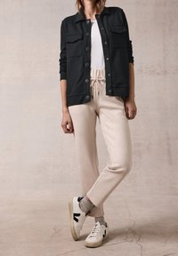 Sort skjortejakke med to brystlommer, båret over en hvid skjorte. Lyse beige sweatpants med justerbar talje og manchetter ved anklerne. Hvide sneakers.