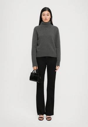Femme portant un pull à col roulé gris, un pantalon noir évasé, des talons ouverts noirs et tenant un petit sac à main noir sur fond blanc.