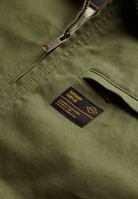 Groene stoffen jas met een zilveren rits en een zwart label met oranje tekst, dat het merk en de verzorgingsinstructies aangeeft.