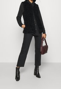 Gilet nero in tessuto peloso sopra una shirt scura, abbinato a pantaloni neri e stivaletti. Gli accessori includono una piccola borsa di un profondo bordeaux.