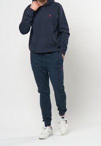 Tmavě modrá mikina s kapucí s nastavitelnými šňůrkami a přední kapsou, v kombinaci s odpovídajícími tepláky s elastickým pasem a logem. Bílé tenisky.