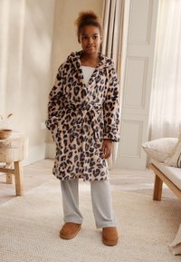 Accappatoio con stampa leopardata in beige e marrone scuro, con cappuccio e cintura annodata. Abbinato a pantaloni grigi e pantofole marroni su un tappeto con trama.