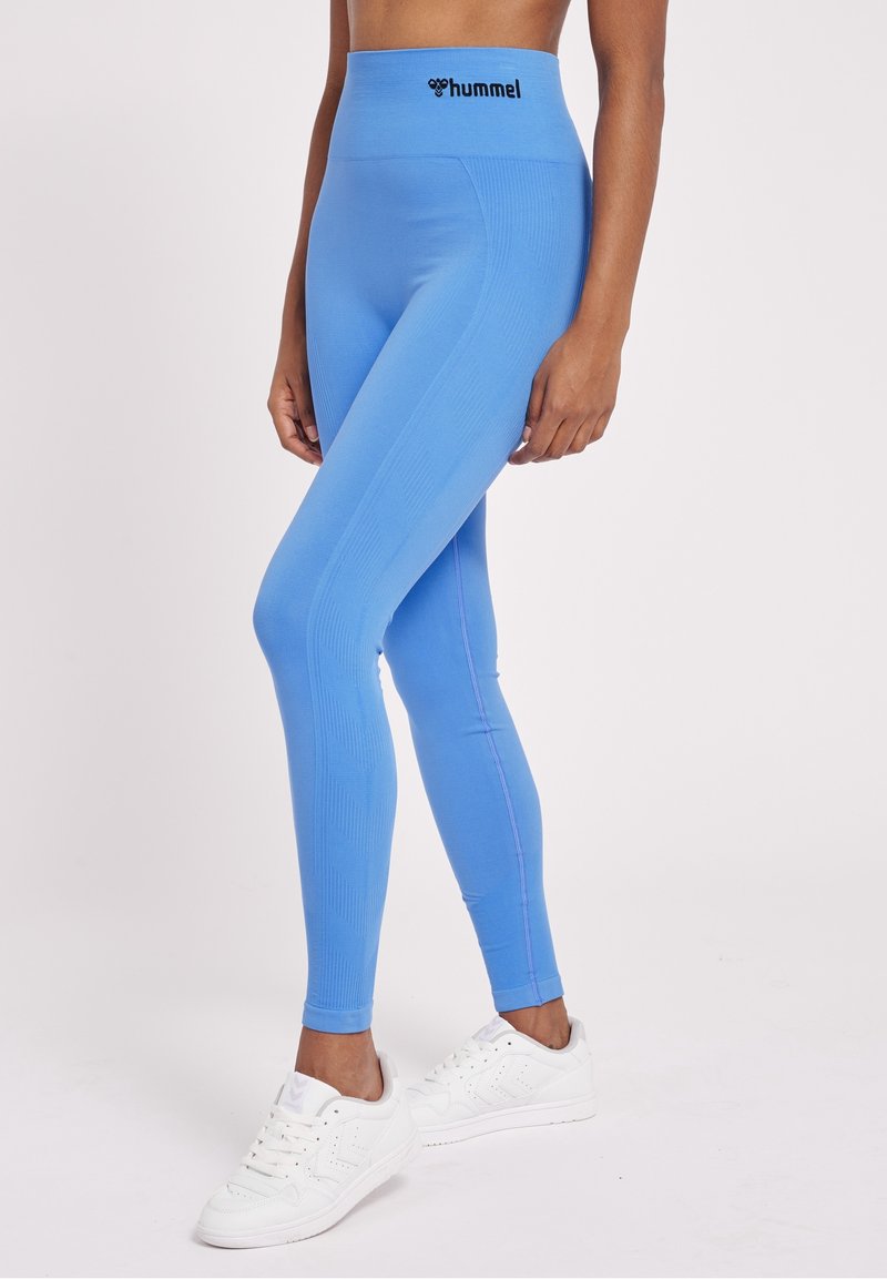 Blå högmidjade leggings tillverkade av stretchigt material, med en texturerad design och kontrasterande rosa sömmar, i kombination med vita sneakers.