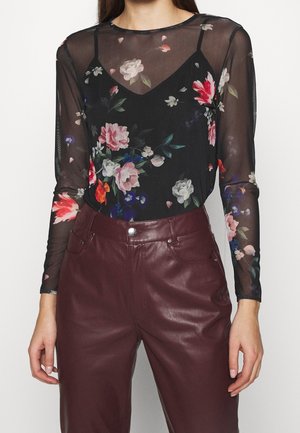 Kobieta ubrana w czarną, przejrzystą, długorękawową bluzkę w kwiaty z czarnym topem na ramiączkach pod spodem oraz wysokowasowe, bordowe skórzane spodnie.