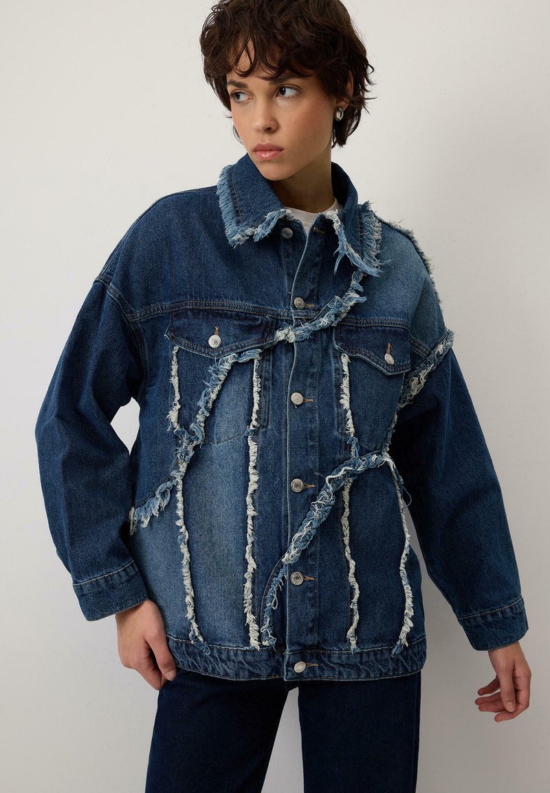 Jauns cilvēks, tērpies tumši zilā applēstā džinsu jakā ar nolietotām malām, stāv pie vienkāršas gaišas sienas, skatoties uz sāniem.