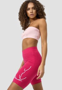 Růžový zkrácený bandeau top v kombinaci s růžovými šortkami s vysokým pasem, na kterých je design bílé abstraktní tváře. Bavlněný materiál, hladká textura.