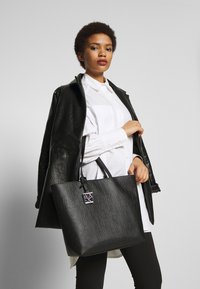 Svart tote bag med preget logo-design, glatt tekstur og doble håndtak; båret med en svart lærjakke og hvit skjorte.