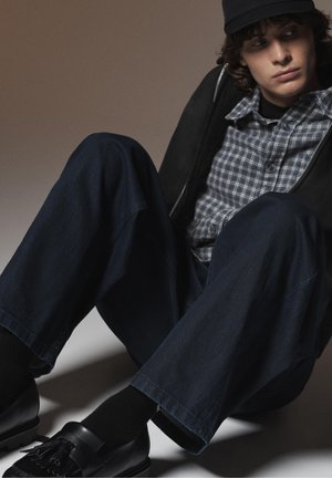 Young man seated on floor wearing black cap, black jacket, gray checkered shirt, dark jeans, black socks, and black tassel loafers.
