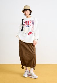 Balta sporta krekls ar izteiksmīgiem sarkaniem un ziliem Nike grafikiem, brūna svārku ar elastīgu apakšu, baltas zeķes un pelēki Nike kedas ar rozā akcentiem.