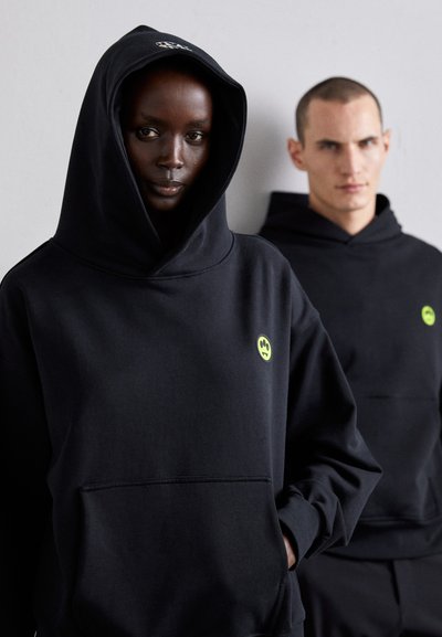 Black hoodies made of soft fabric, featuring a large hood and a small neon yellow logo on the chest. Front pocket design adds functionality.