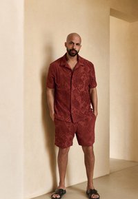 Maglietta a maniche corte color marrone scuro e pantaloncini abbinati con un design texturizzato e decorato. Abbinati a sandali scuri su uno sfondo beige neutro.