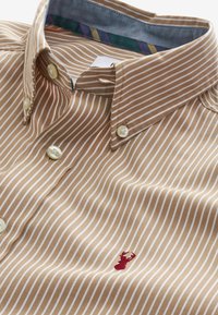 Chemise à rayures beige et blanche avec col boutonné et petite broderie rouge de homard sur la poitrine.