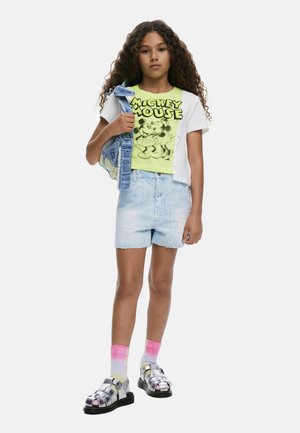 Grafikus póló neon sárga és fehér mintával, világoskék farmer rövidnadrág, ezüst szandál és rózsaszín ombre zokni. Farmer dzseki a karra terítve.