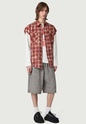 Giovane che indossa una camicia a quadri rossa senza maniche sopra una maglietta bianca a maniche lunghe, pantaloncini larghi grigi, calzini bianchi e scarpe nere con lacci.