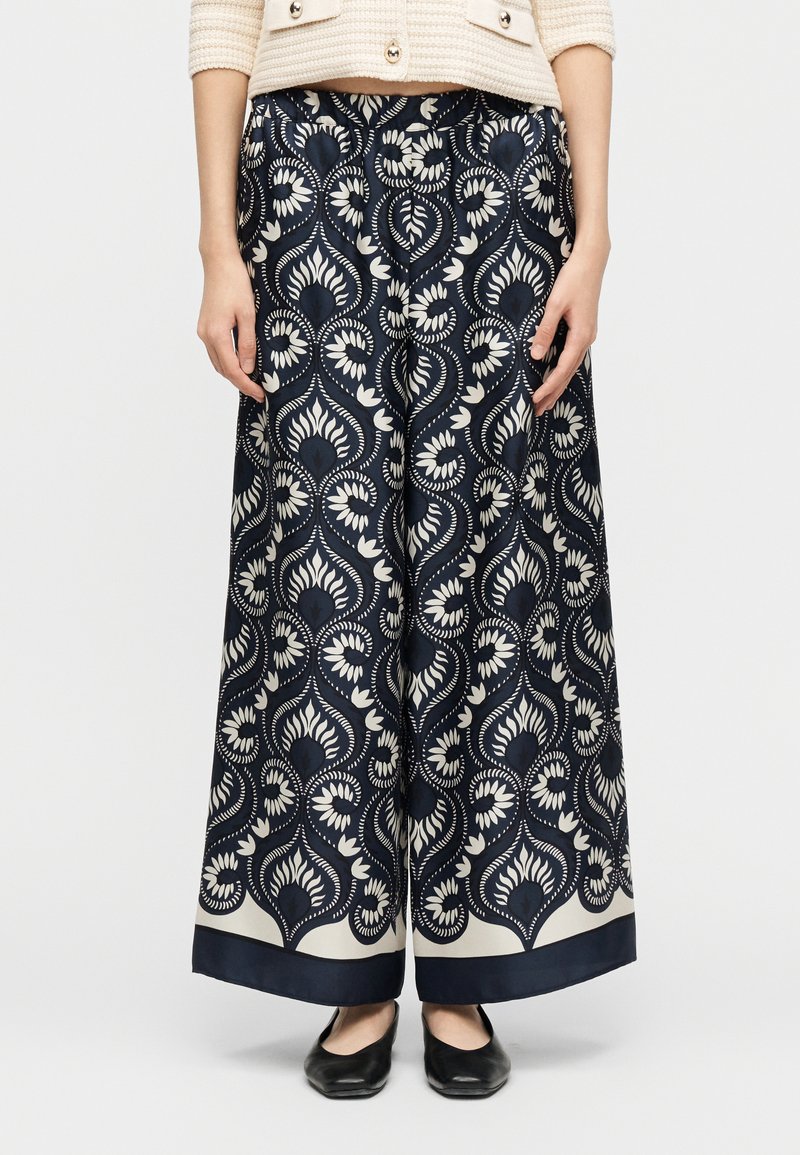 Φαρδιά παντελόνια σε ναυτικό μπλε με λευκά ανθισμένα και γεωμετρικά σχέδια, συνδυασμένα με μαύρα μπαλαρίνες και ένα μπεζ υφασμάτινο σακάκι.